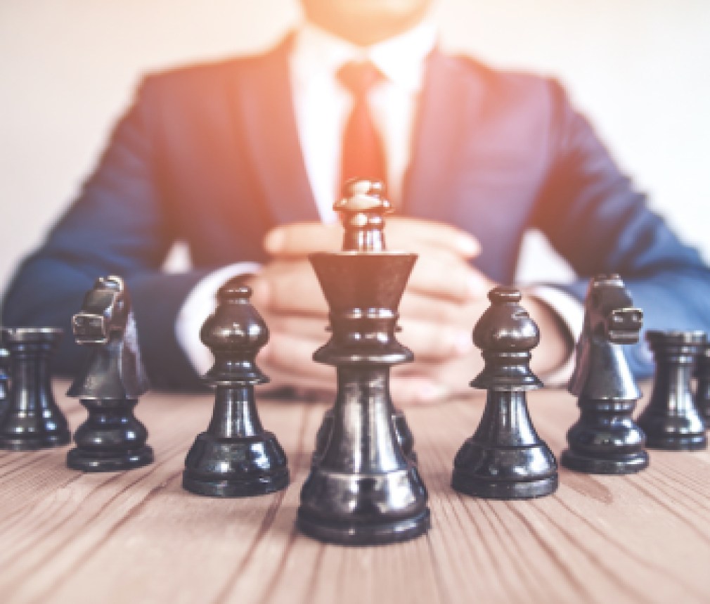 Retro style image of a businessman with clasped hands planning strategy with chess figures on an old wooden table.