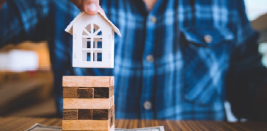 Hand holding a piece of wood block with model white house on dollar banknote. Insurance and property investment real estate concept.