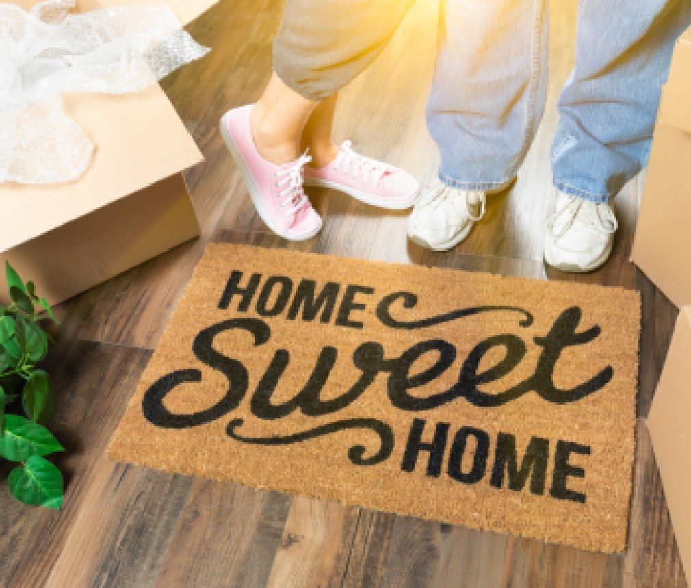 Man and Woman Standing Near Home Sweet Home Welcome Mat, Moving Boxes and Plant.