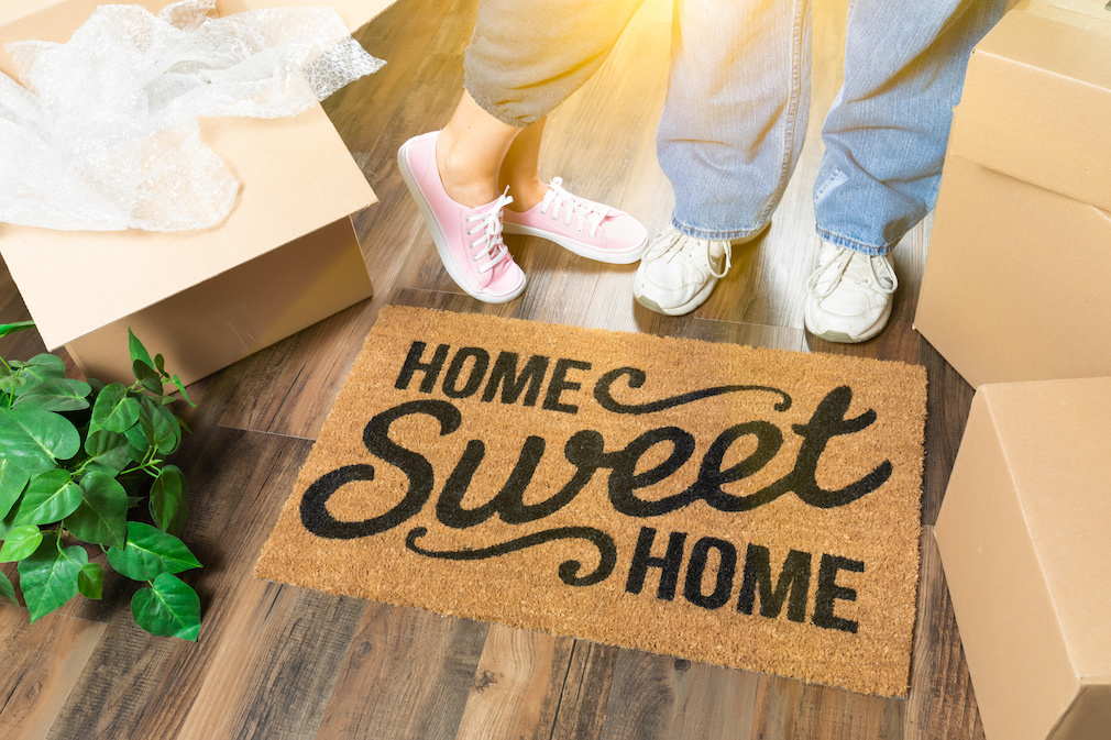 Man and Woman Standing Near Home Sweet Home Welcome Mat, Moving Boxes and Plant.