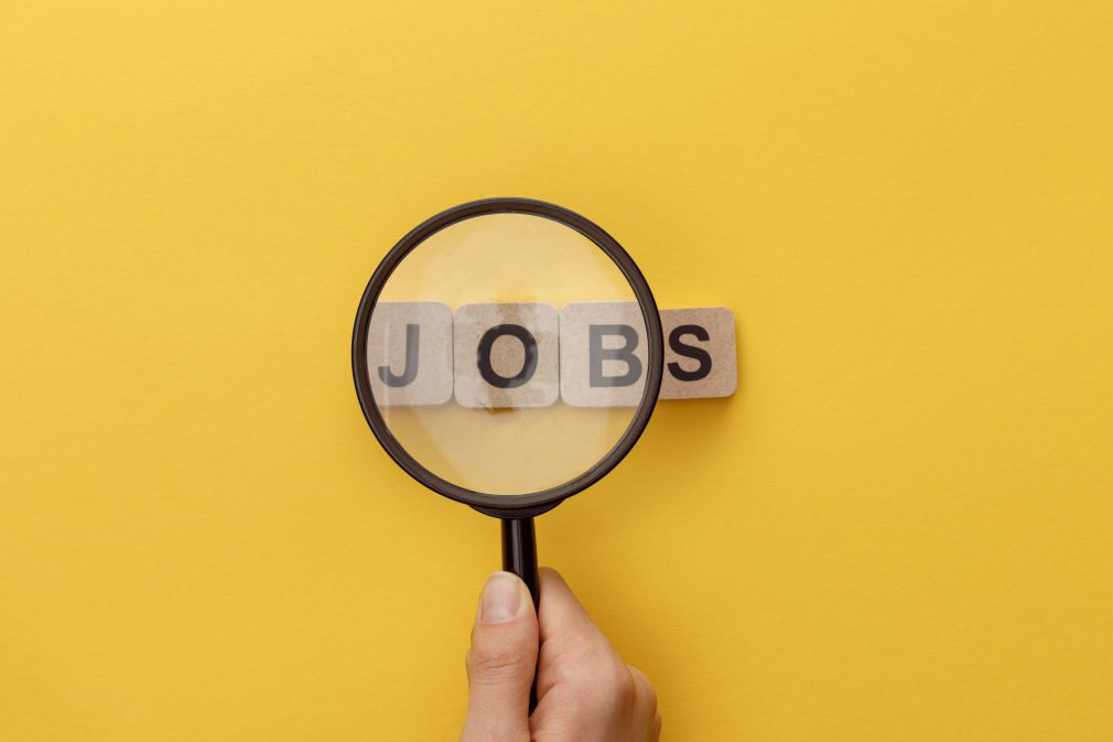 cropped view of woman holding magnifying glass under cardboard squares with jobs lettering on yellow background