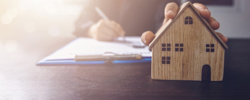real estate, property and home owner signing contract concept, small wooden house model on office table with hand of woman buyer sign on rental agreement paper to rent above mentioned residence