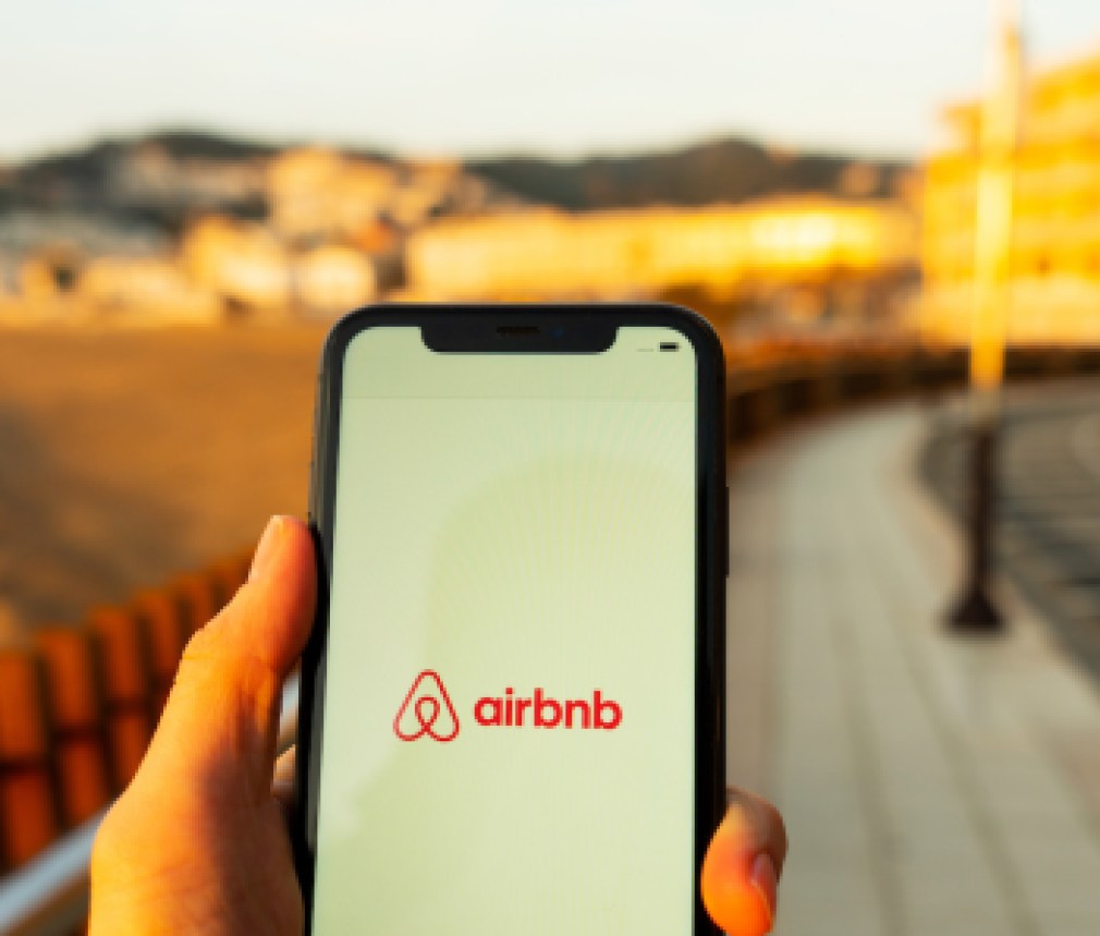 Hands holding iPhone 11 with an app of airbnb service. Holidays and vacation concept. A woman finding a place to stay or booking a hotel, apartment on a sunny day next to Mediterranean sea