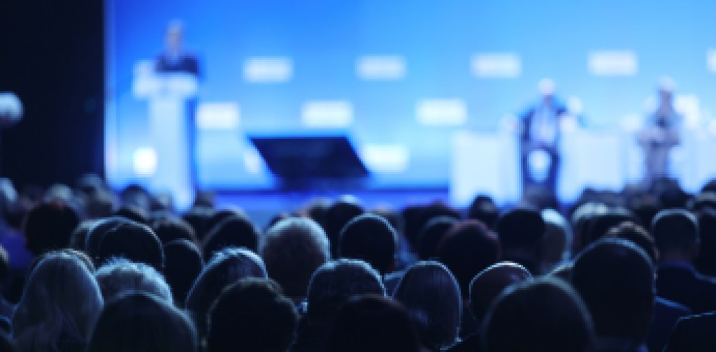 Business and entrepreneurship symposium. Speaker giving a talk at business meeting. Audience in the conference hall. Rear view of unrecognized participant in audience.