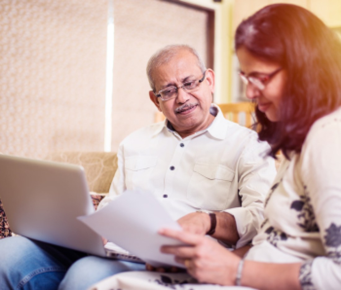 Senior Indian/asian couple accounting, doing home finance and checking bills with laptop, calculator and money while sitting on sofa/couch at home