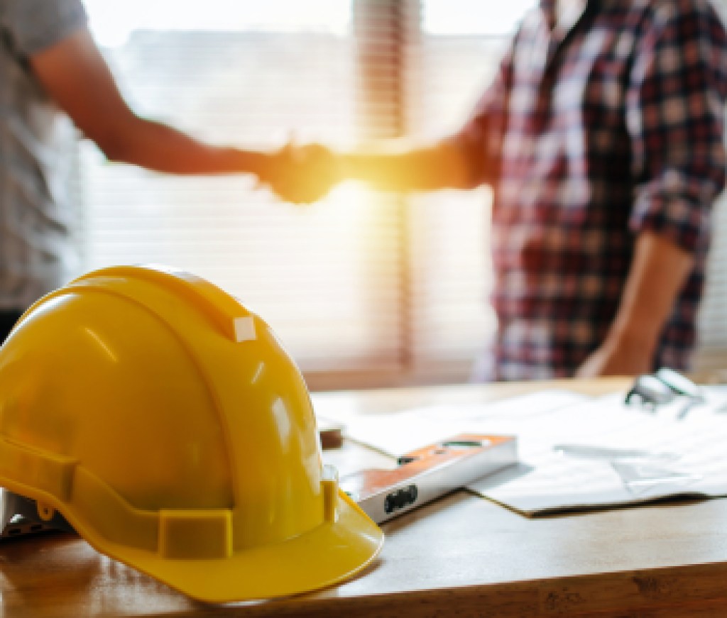 yellow safety helmet on workplace desk with construction worker team hands shaking greeting start up plan new project contract in office center at construction site, partnership and contractor concept