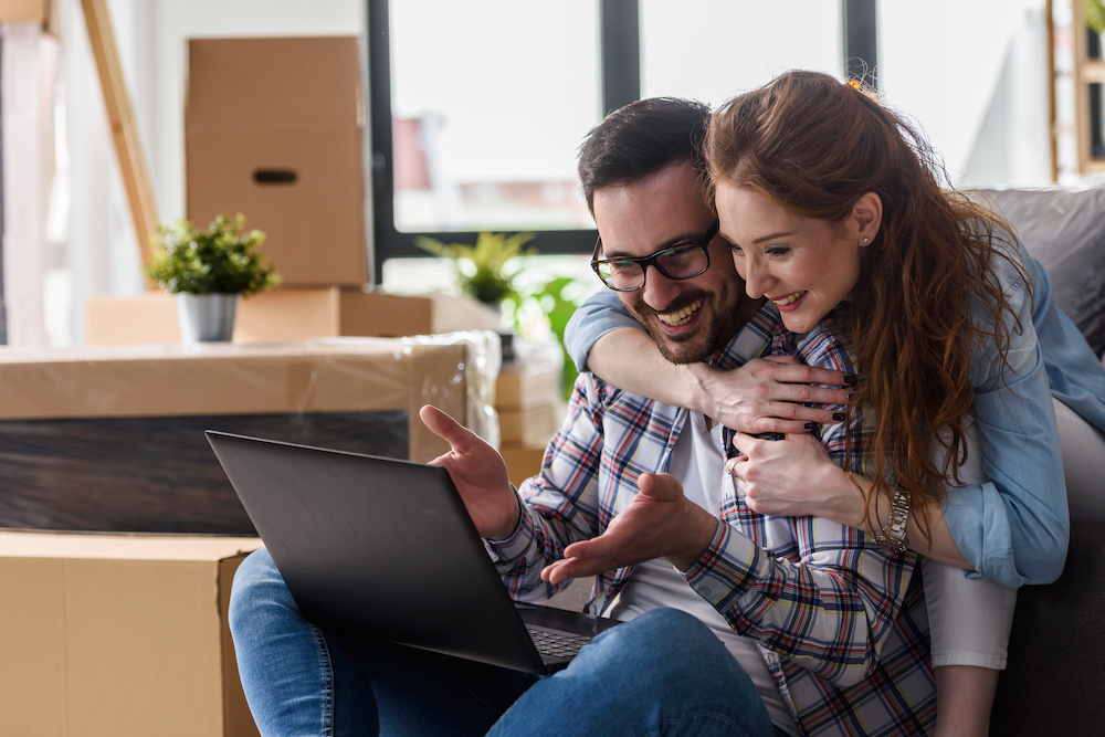 Young couple moving in new home. Sitting and relaxing after unpacking. Searching home decorating ideas on laptop