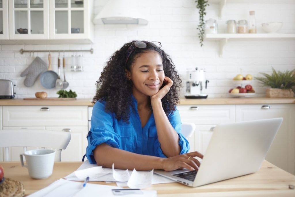 Indoor shot of happy young Afro American woman using portable computer and smiling, enjoying modern technology while paying bills for rent, gas and electricity online, sitting at kitchen table