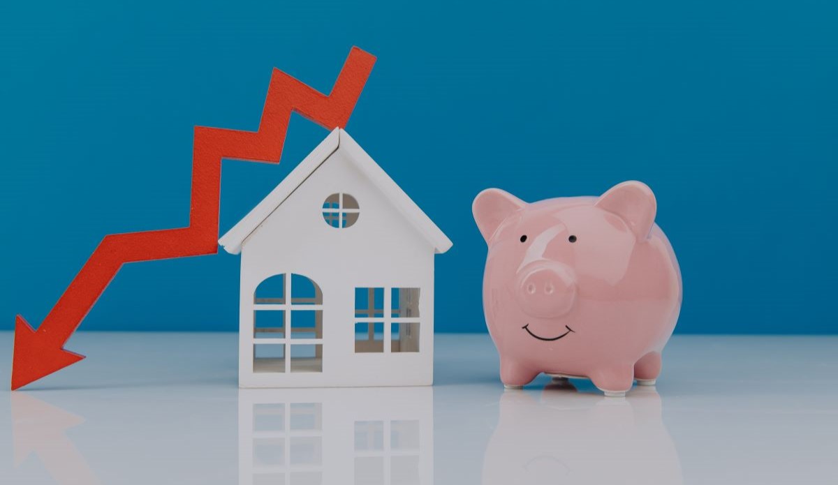 smiling piggy bank next to model home and arrow pointing down mortgage rates decrease concept